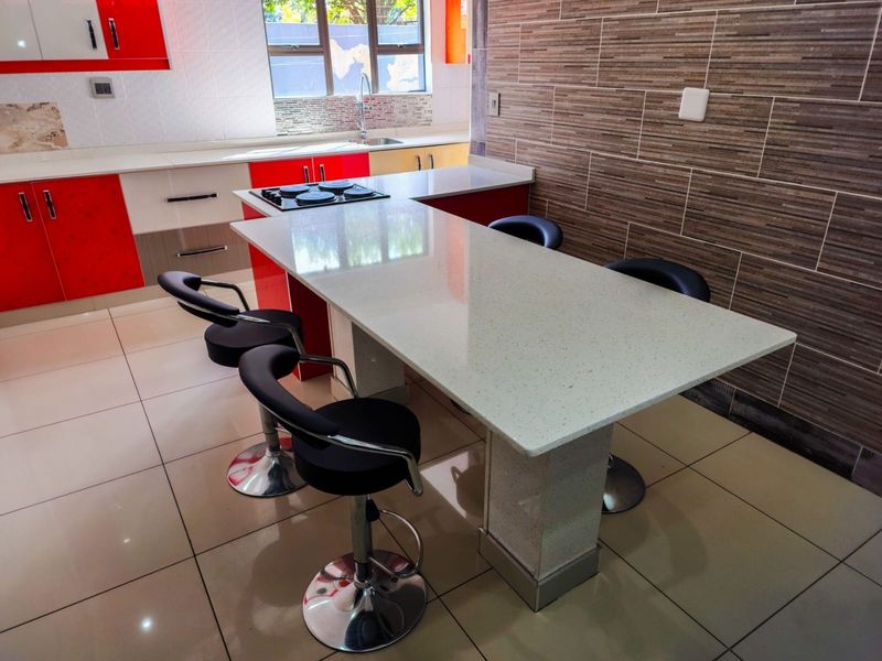 Modern kitchen featuring a central quartz breakfast bar, built-in hob, high-gloss cabinetry, and contemporary tiled feature wall with neutral floor tiles.