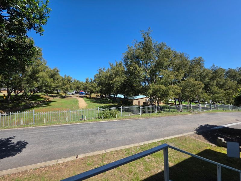 A view of the street and surrounding neighborhood area, featuring established trees, open green space, and secure metal perimeter fencing.