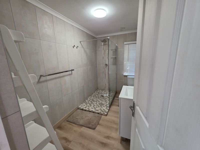 A modern bathroom featuring a walk-in shower with glass screen, vanity basin, toilet, and neutral wall tiling with wood-look flooring.