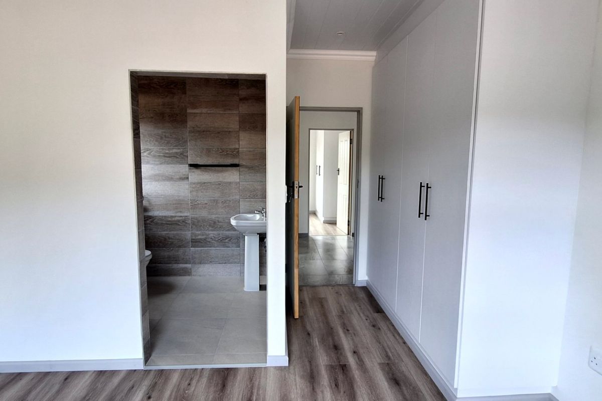 A bright, empty bedroom featuring modern laminate flooring, built-in cupboards, and direct access to a tiled en-suite bathroom with a pedestal basin.