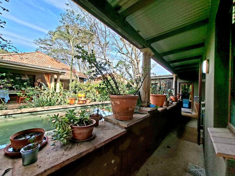 A covered patio area featuring a stone ledge, classic columns, and a corrugated roof, overlooking a garden and water feature.