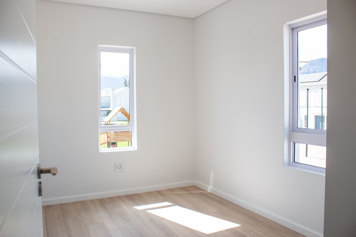 A bright, empty bedroom featuring modern laminate flooring, clean white walls, and large aluminium-framed windows allowing for ample natural light.