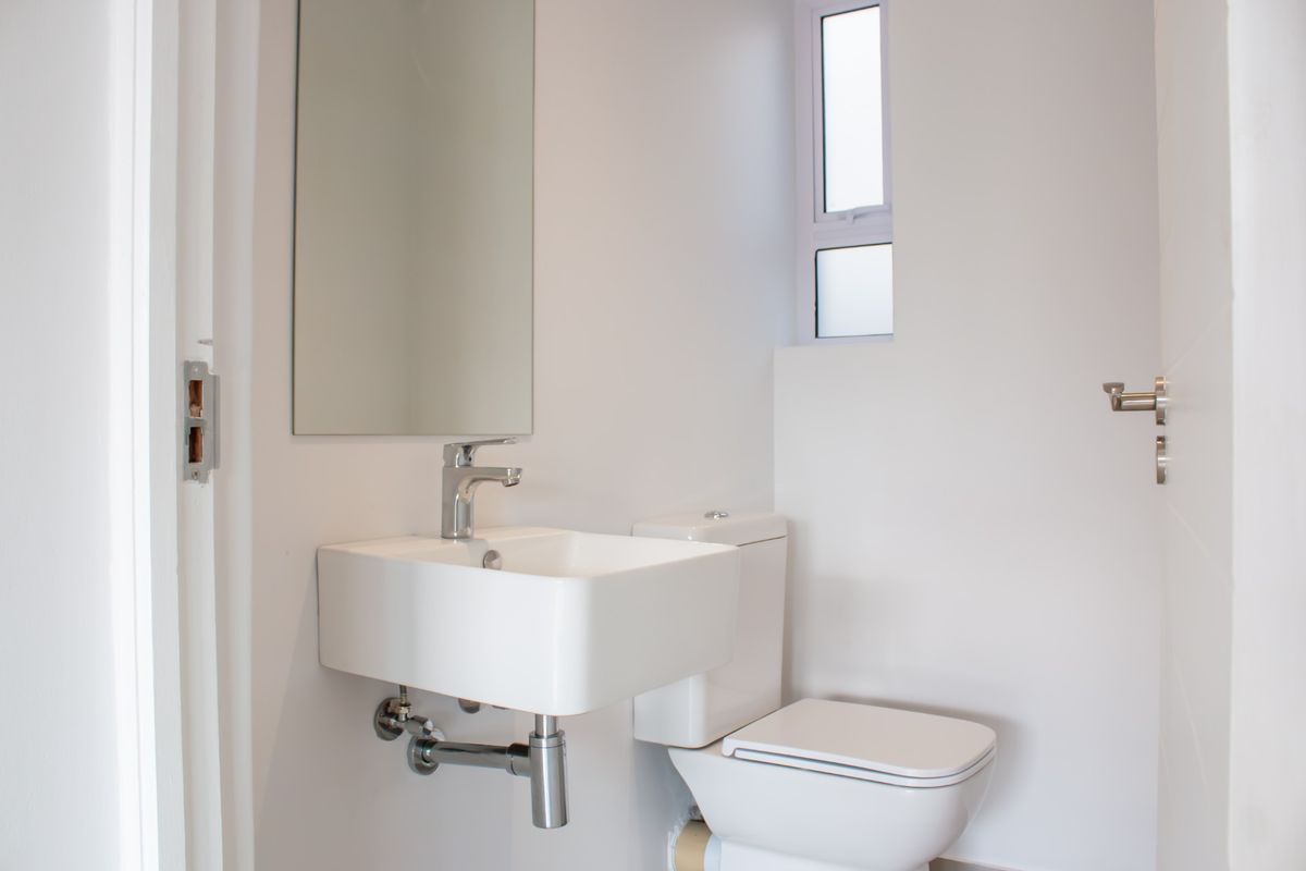 A clean, modern guest bathroom featuring a wall-mounted basin, chrome fixtures, and a toilet, finished with neutral white walls and a frosted window.