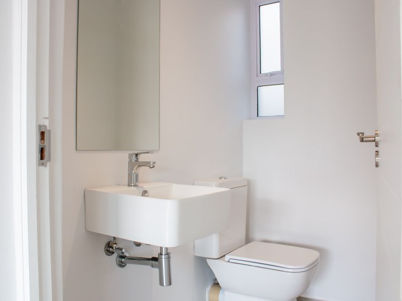 A clean, modern guest bathroom featuring a wall-mounted basin, chrome fixtures, and a toilet, finished with neutral white walls and a frosted window.
