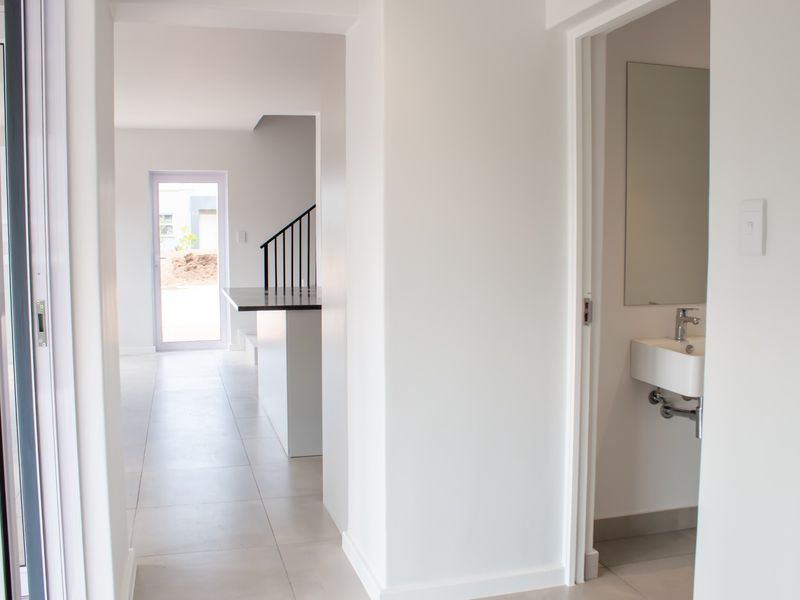 A bright, modern hallway featuring neutral tiled flooring, a minimalist wall-mounted basin, and a view through to the staircase and rear entrance.