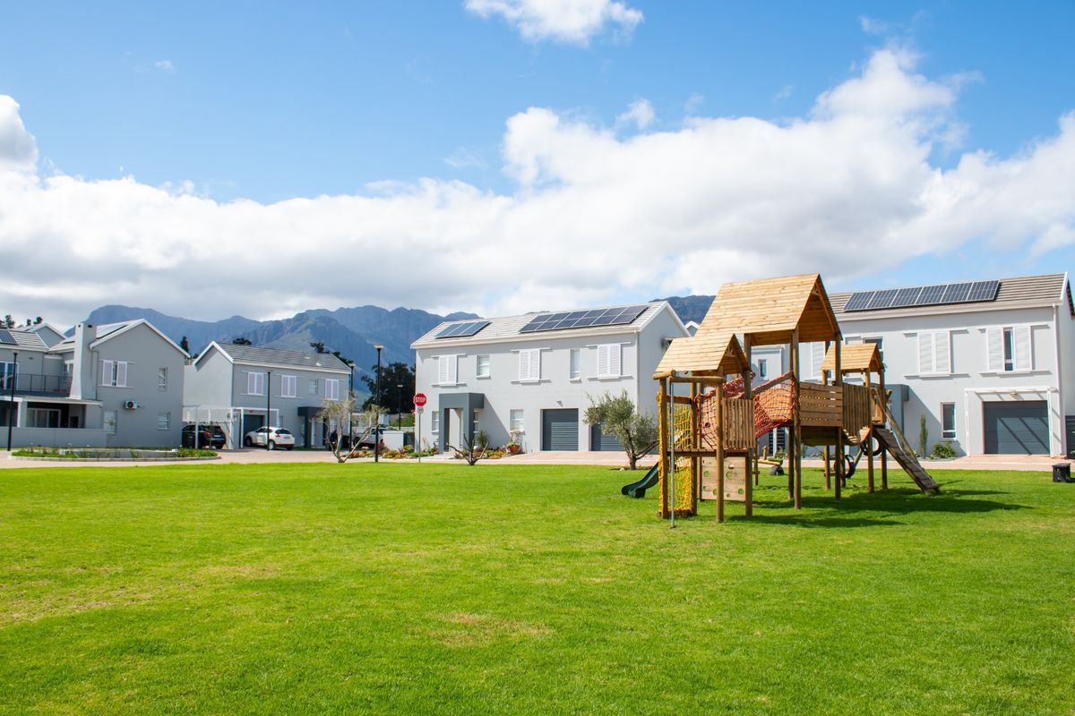 A well-maintained residential complex featuring modern townhouses with solar panels, paved driveways, and a communal green space with a playground.