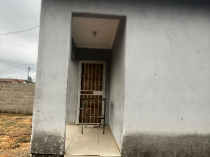 Front entrance of a house featuring a security gate, tiled porch area, and concrete apron, showing signs of exterior wall weathering and damp.