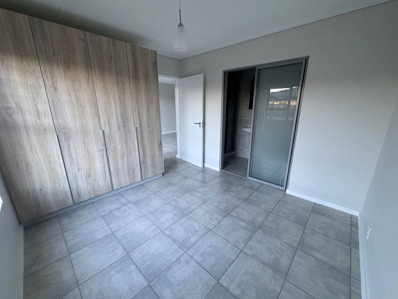 A modern, empty bedroom featuring neutral tiled flooring, extensive built-in wooden cupboards, and a sliding door leading to an en-suite bathroom.