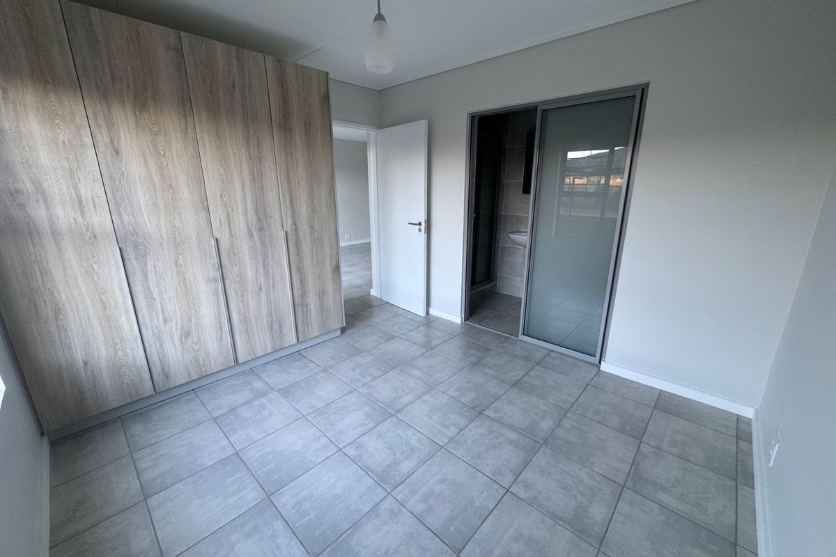 A modern, empty bedroom featuring neutral tiled flooring, extensive built-in wooden cupboards, and a sliding door leading to an en-suite bathroom.