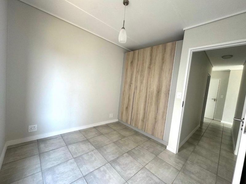 A clean, empty bedroom featuring modern wood-finish built-in cupboards, neutral tiled flooring, and a simple pendant light fixture.