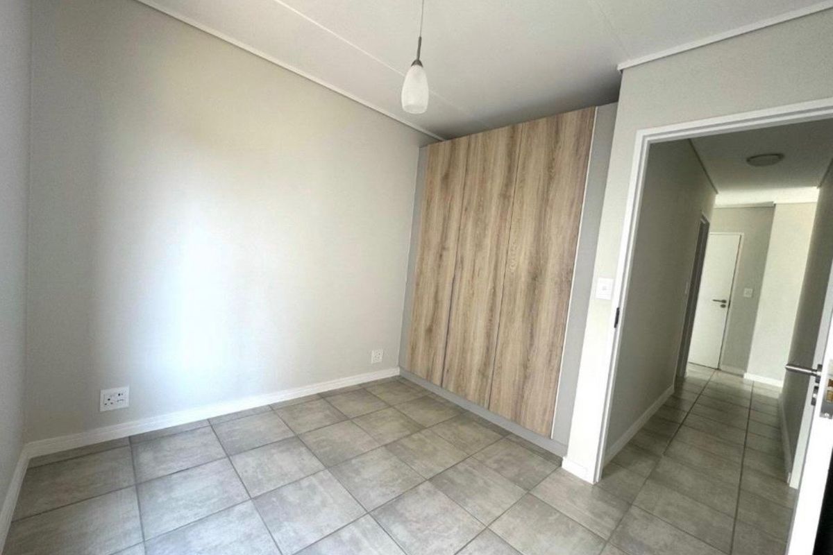 A clean, empty bedroom featuring modern wood-finish built-in cupboards, neutral tiled flooring, and a simple pendant light fixture.