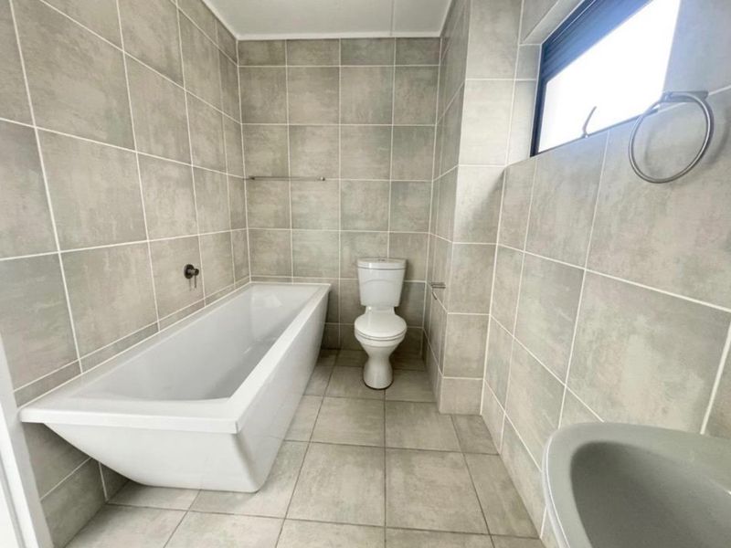 A clean, modern bathroom featuring neutral grey wall and floor tiles, a white bathtub, and a standard toilet.