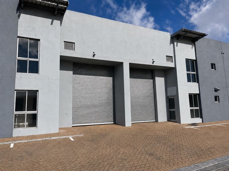 Modern industrial warehouse facade featuring dual roller shutter doors, paved forecourt, and contemporary grey exterior finish with aluminium window frames.