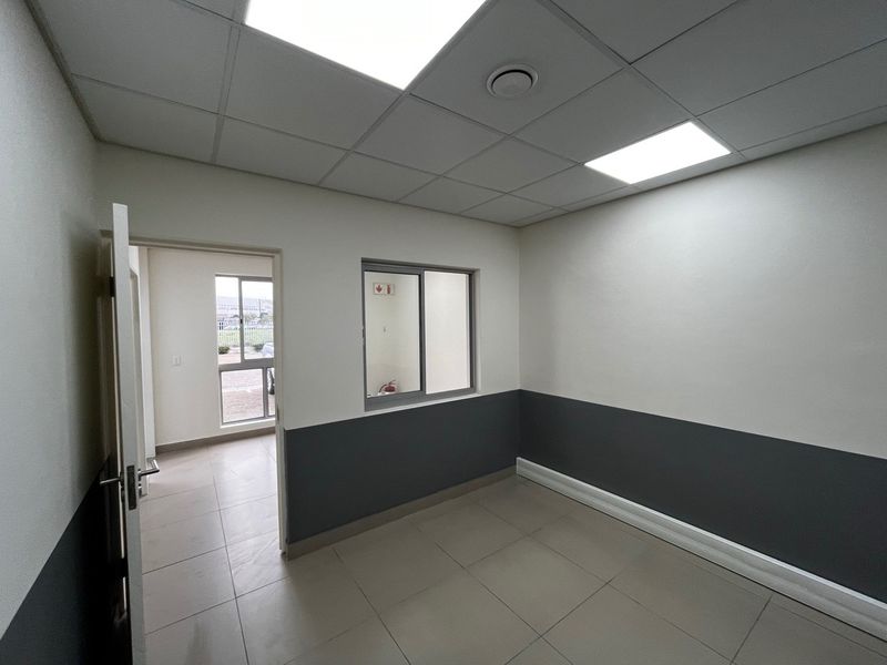 A clean, empty office space featuring tiled flooring, a suspended ceiling with recessed lighting, and neutral wall finishes with a dark grey dado.