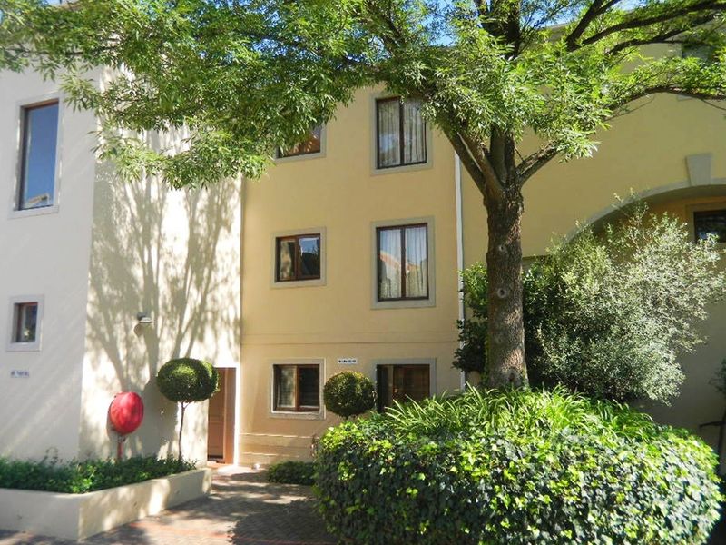 Front elevation of the apartment building featuring a paved entrance area, established trees, and landscaped garden beds.