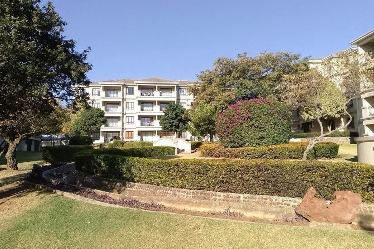 A view of the apartment complex exterior featuring well-maintained landscaped gardens, manicured hedges, and a paved walkway leading to the building.