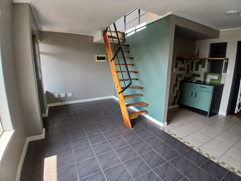 Open plan living area featuring a wooden staircase, tiled flooring, and a compact kitchen corner with cabinetry and tiled backsplash.