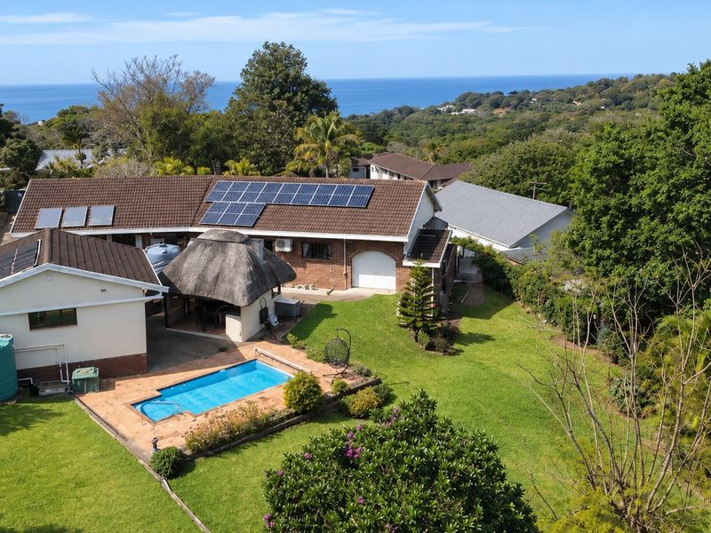 Aerial view of a residential property featuring a swimming pool, thatched lapa, solar panels, and a spacious garden with ocean views.