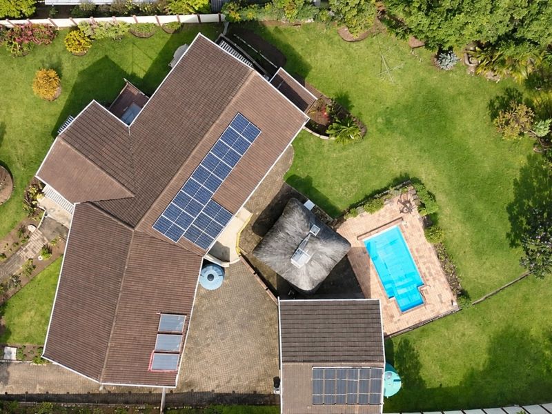 Aerial view of the property featuring a large garden, swimming pool, thatched lapa, paved driveway, and solar panel installations on the roof.