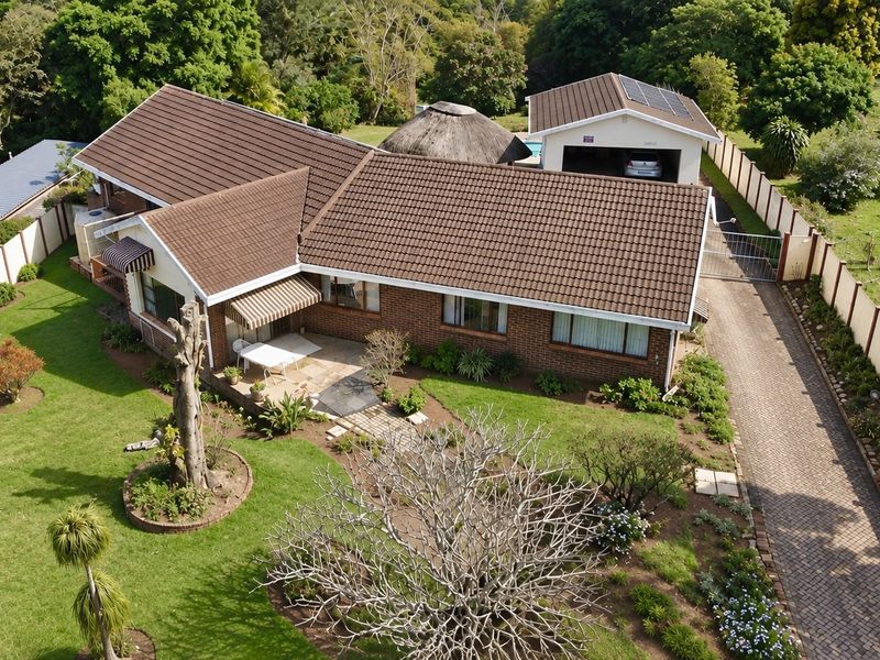 Aerial view of a brick-faced house featuring a tiled roof, paved driveway, detached garage with solar panels, and a spacious landscaped garden.