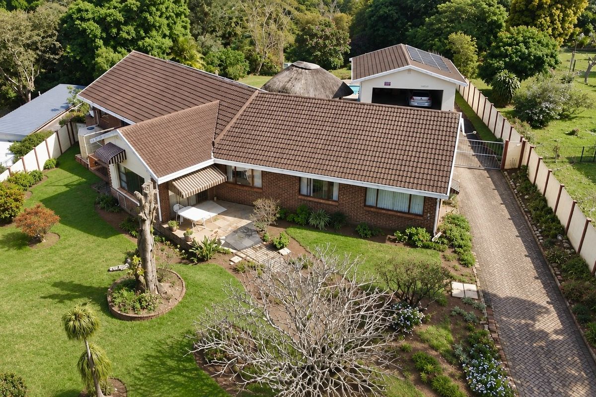 Aerial view of a brick-faced house featuring a tiled roof, paved driveway, detached garage with solar panels, and a spacious landscaped garden.