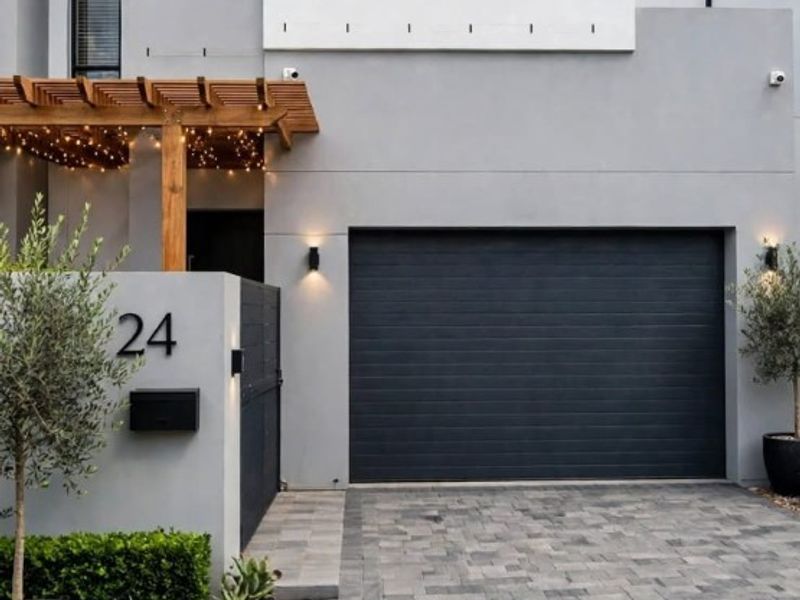 Modern two-story residential facade featuring a sleek grey finish, a dark sectional garage door, and a timber pergola over the entrance.