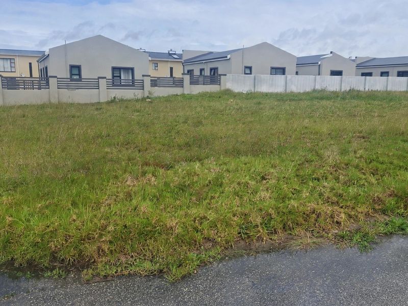 A vacant residential plot of land featuring grass cover, situated in a developed neighborhood with established boundary walls and road access.
