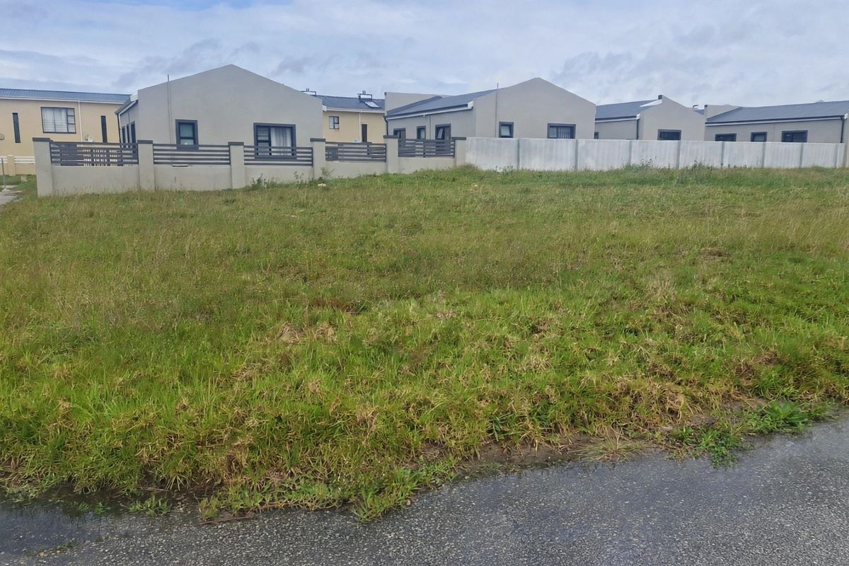 A vacant residential plot of land featuring grass cover, situated in a developed neighborhood with established boundary walls and road access.