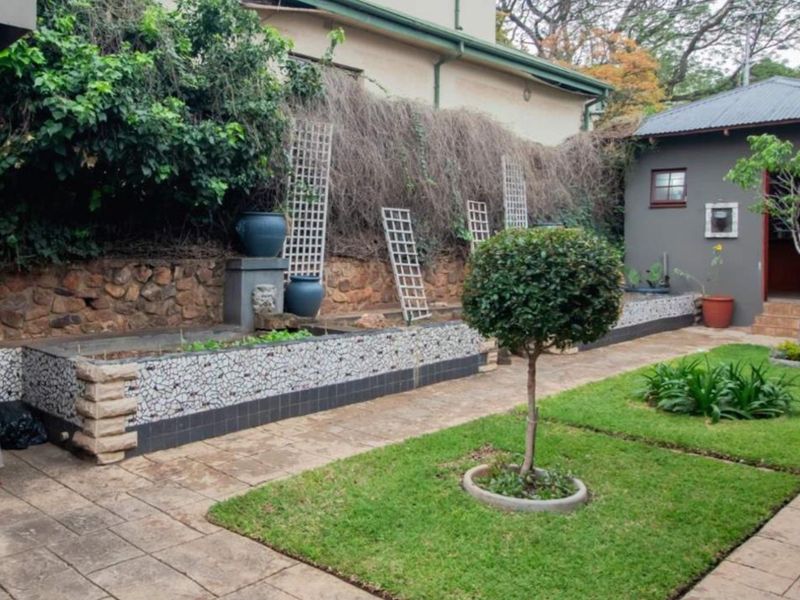 A well-maintained rear garden featuring a stone retaining wall, mosaic-tiled planter boxes, paved pathways, and a neat lawn area with an adjacent outbuilding.