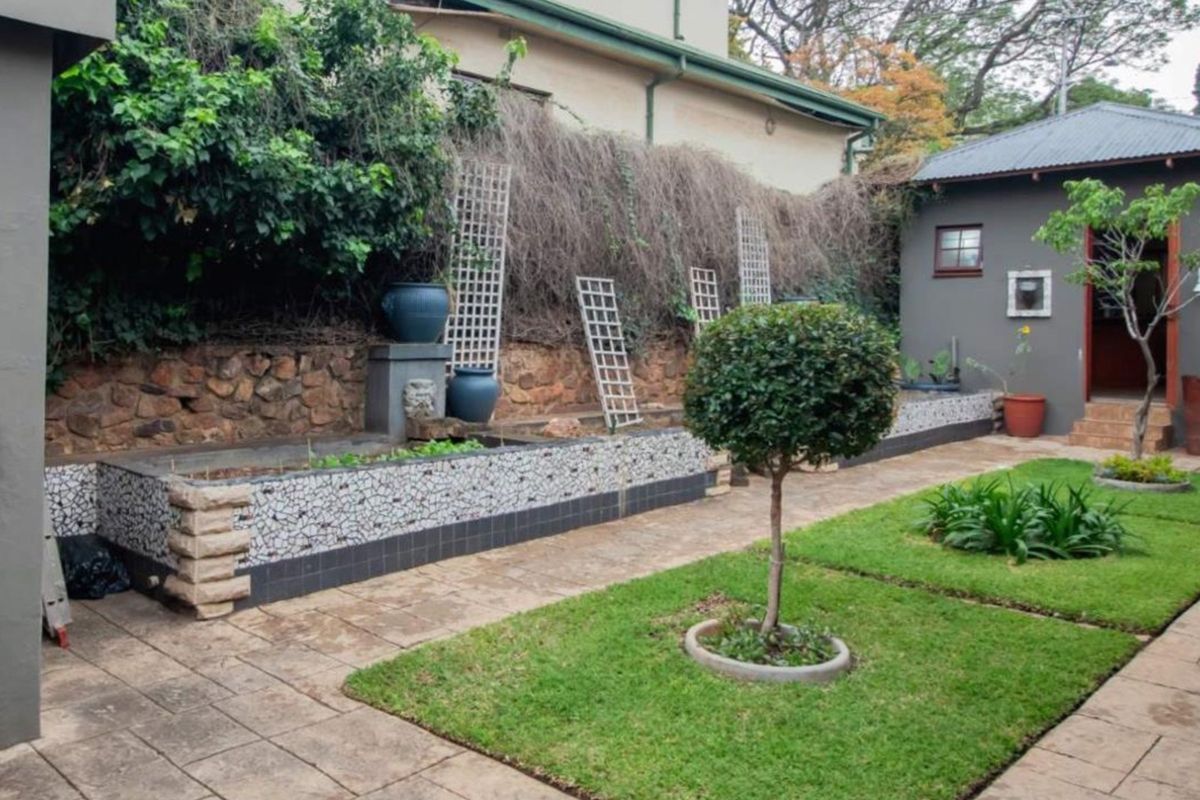 A well-maintained rear garden featuring a stone retaining wall, mosaic-tiled planter boxes, paved pathways, and a neat lawn area with an adjacent outbuilding.