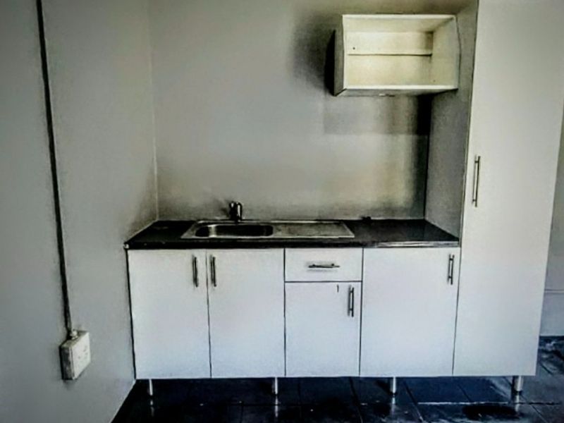 A compact kitchen area featuring white built-in cabinetry, a stainless steel sink, and dark tiled flooring, presenting a functional but dated utility space.