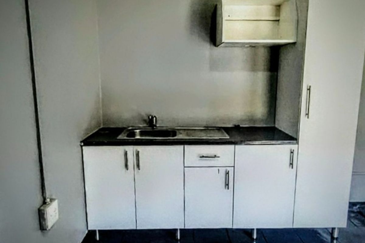 A compact kitchen area featuring white built-in cabinetry, a stainless steel sink, and dark tiled flooring, presenting a functional but dated utility space.