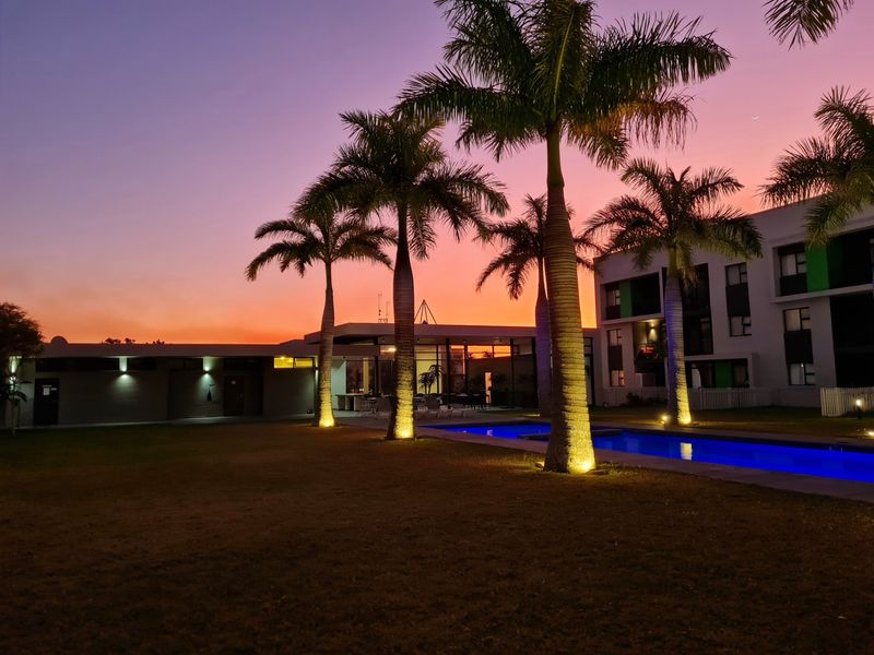 A beautifully lit outdoor pool area at sunset, featuring mature palm trees and modern apartment building architecture.