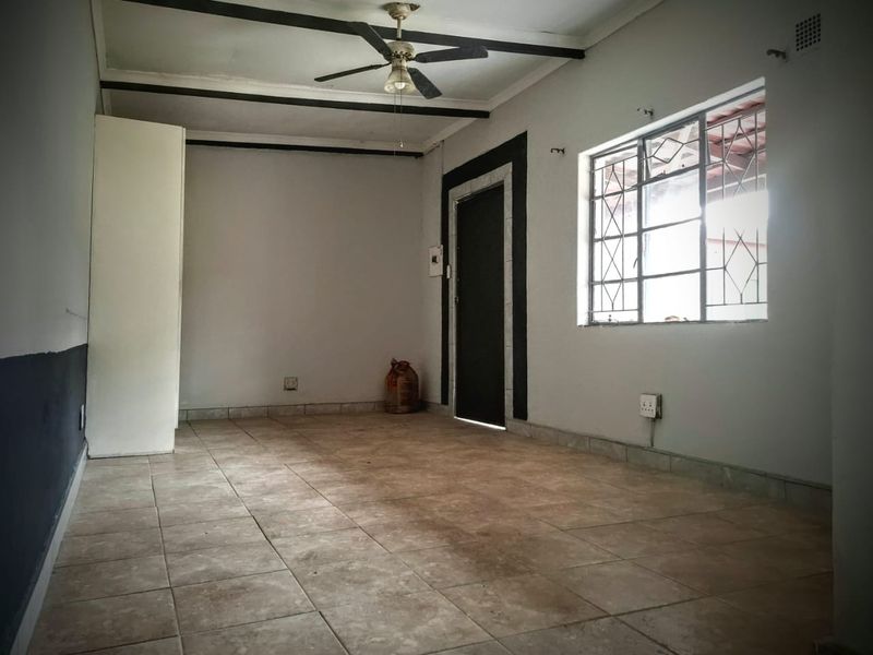 An empty living room featuring tiled flooring, exposed ceiling beams, a ceiling fan, and security bars on the window.