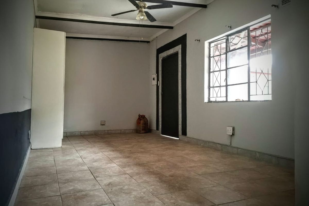 An empty living room featuring tiled flooring, exposed ceiling beams, a ceiling fan, and security bars on the window.