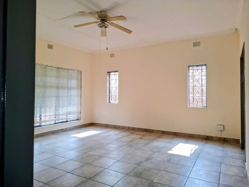An empty living room featuring tiled flooring, a ceiling fan, and windows fitted with security bars and venetian blinds.