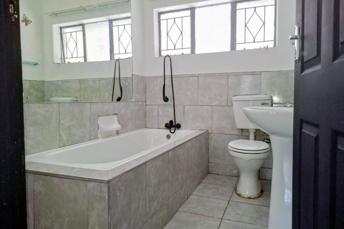 A functional bathroom featuring a built-in bathtub, pedestal sink, toilet, and tiled finishes, complemented by natural light from windows with security bars.