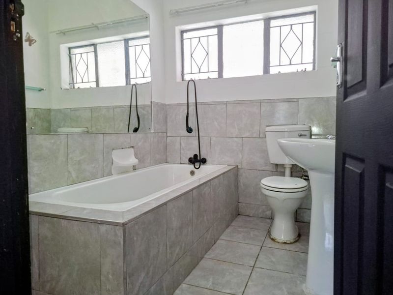 A functional bathroom featuring a built-in bathtub, pedestal sink, toilet, and tiled finishes, complemented by natural light from windows with security bars.