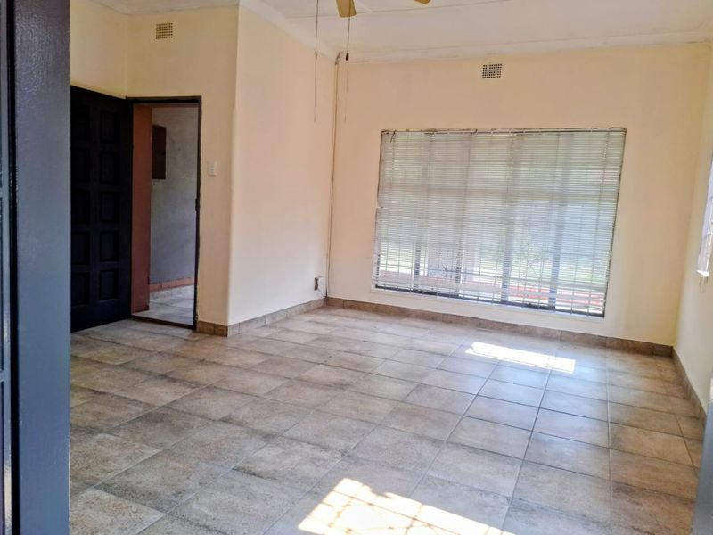 A vacant living room featuring neutral tiled flooring, a ceiling fan, and large windows fitted with venetian blinds.