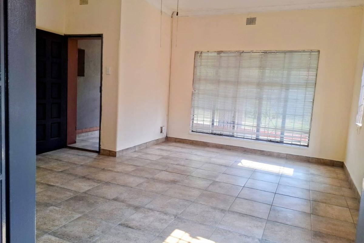 A vacant living room featuring neutral tiled flooring, a ceiling fan, and large windows fitted with venetian blinds.