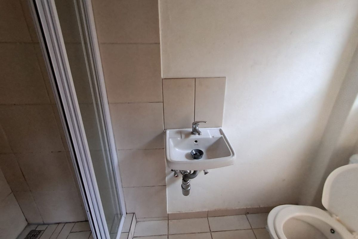A compact bathroom featuring a wall-mounted basin, shower enclosure, and toilet with tiled finishes, requiring maintenance and cosmetic updates.