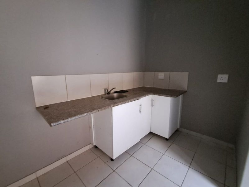 A compact, empty kitchen area featuring white built-in cabinetry, a laminate countertop with an integrated stainless steel sink, and tiled flooring.