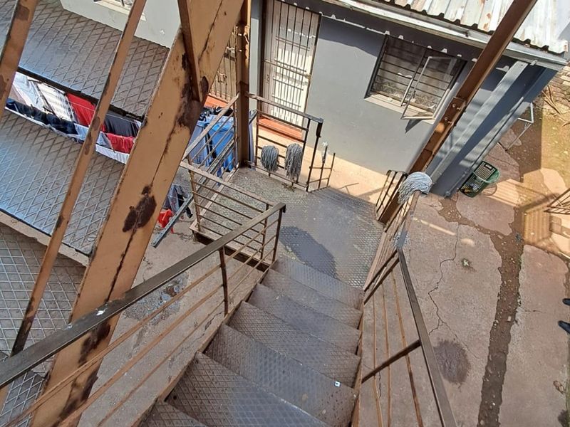 An elevated view of an exterior rear area featuring a weathered metal staircase, security-fitted windows, and a corrugated metal roof structure.