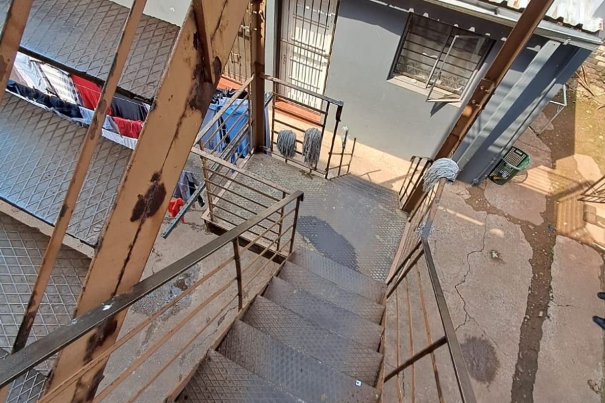 An elevated view of an exterior rear area featuring a weathered metal staircase, security-fitted windows, and a corrugated metal roof structure.
