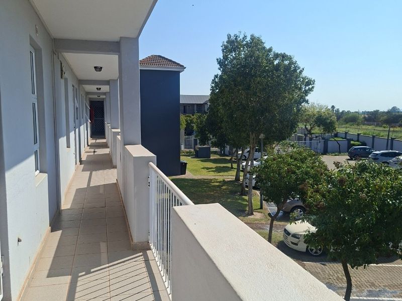 A covered exterior balcony walkway featuring tiled flooring and a metal balustrade, overlooking the apartment complex grounds and parking area.