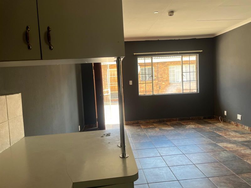 Open plan living area featuring tiled flooring, kitchen cabinetry with a breakfast counter, and natural light through a window with security bars.