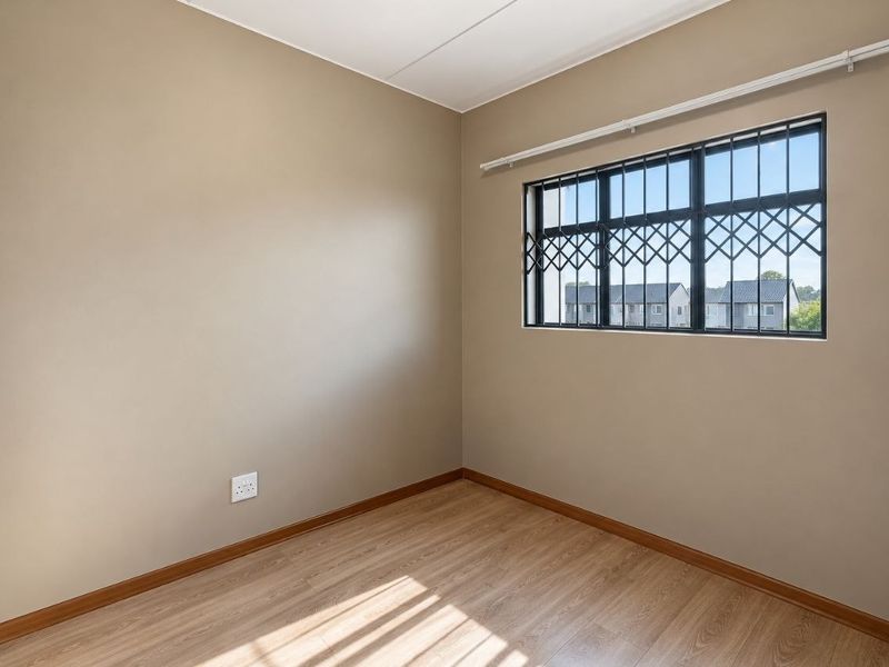 A clean, empty bedroom featuring light-toned laminate flooring, neutral walls, and a window fitted with security bars and a curtain rail.