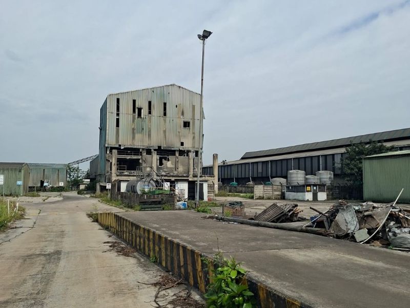 Industrial site featuring a large concrete loading bay, weathered metal-clad structures, and extensive paved yard space in need of significant maintenance.