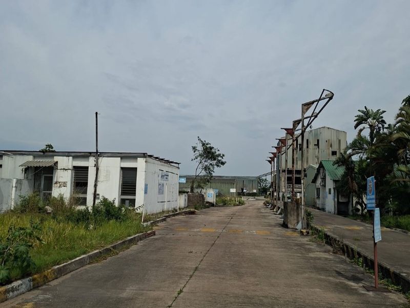 Industrial property exterior featuring a concrete driveway, multiple warehouse structures, and an overhead lighting gantry in a weathered condition.