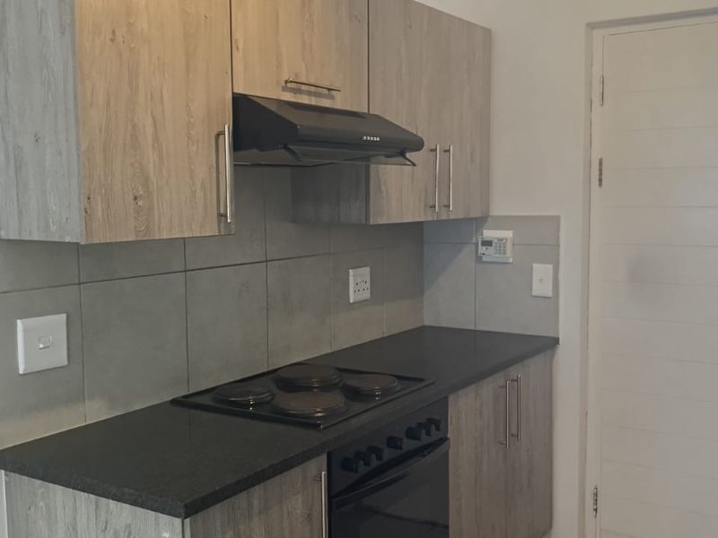 A clean, modern kitchen featuring wood-look cabinetry, dark countertops, a built-in electric hob, oven, and extractor fan with a neutral tiled backsplash.